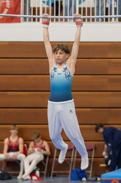 Young male gymnast hangs from the horizontal bar with intense focus, wearing a blue leotard and white trousers.