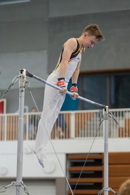 Young male gymnast grips the horizontal bar with intense focus, body horizontal, wearing a black and yellow leotard.