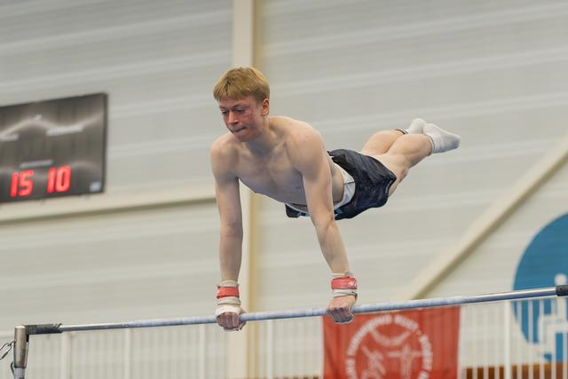 Young male gymnast grips the horizontal bar with intense focus, body extended in a straddle position during indoor competition.