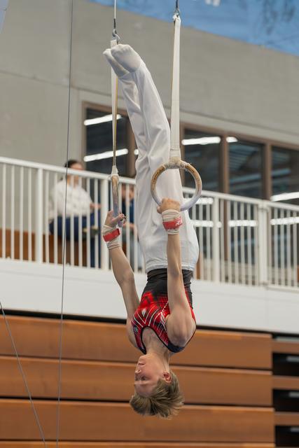 Young male gymnast hangs inverted on still rings, smiling upside-down, wearing red plaid shorts in an indoor gymnasium.