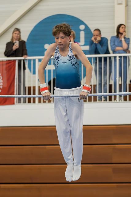 Young male gymnast performing on parallel bars, gripping tightly with focused expression, wearing blue leotard and white trousers.