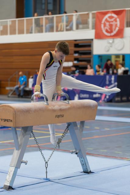 Young male gymnast performs a pommel horse routine, legs extended, with intense focus during an indoor gymnastics meet.