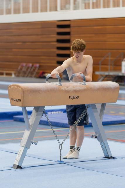 A shirtless teenage gymnast grips the pommel horse handles, focused and preparing his routine in an indoor gymnasium.