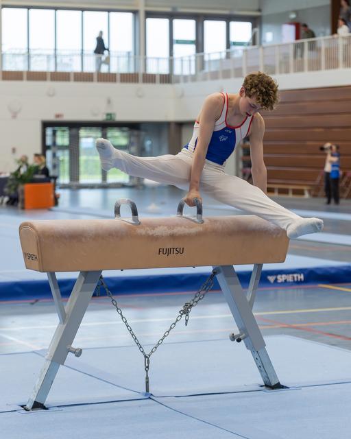 Young male gymnast performs a split on the pommel horse, focused and controlled, gripping the handles firmly indoors.