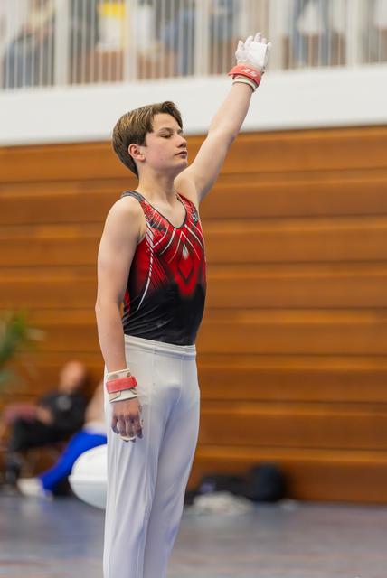 Young male gymnast raises one arm in a salute, eyes closed, poised and focused, wearing a red and black leotard with grips.