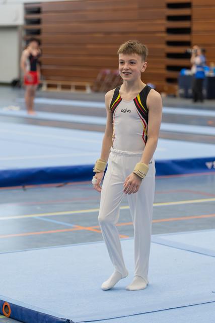 Young male gymnast smiles confidently on the floor mat, wearing white leotard and wrist guards at an indoor gymnastics meet.