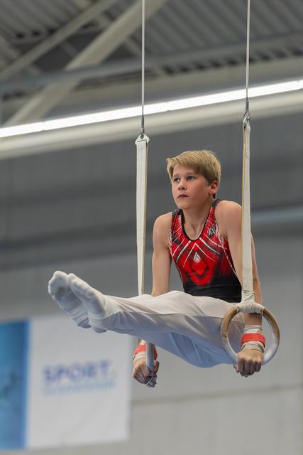 Young male gymnast holds a L-sit on still rings, focused gaze upward, wearing red and black leotard in an indoor gym.