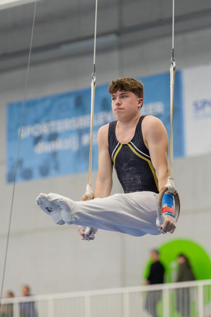 Young male gymnast holds a tuck position on still rings, wearing a navy leotard, with a focused, determined expression.