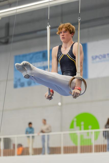 Teenage male gymnast holds an L-sit on still rings, focused expression, competing in an indoor gymnastics hall.