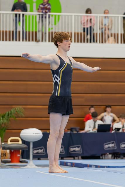 Red-haired teenage gymnast stands poised on the floor mat, arms outstretched with focused concentration during his routine.