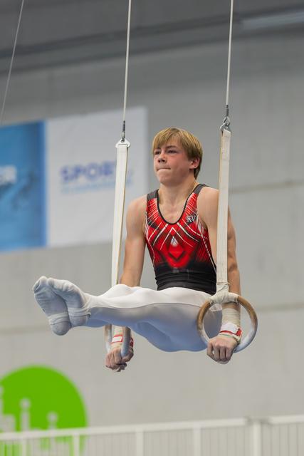 Young male gymnast holds a tuck position on still rings, his focused gaze directed upward during a gymnastics meet.