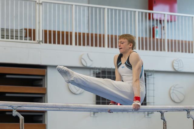 Red-haired teenage gymnast performs a straddle L-sit on parallel bars, focused expression, indoor gymnasium.