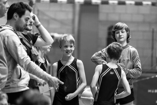 Coach gestures while speaking to four young gymnasts in black leotards, with one athlete facing away from camera