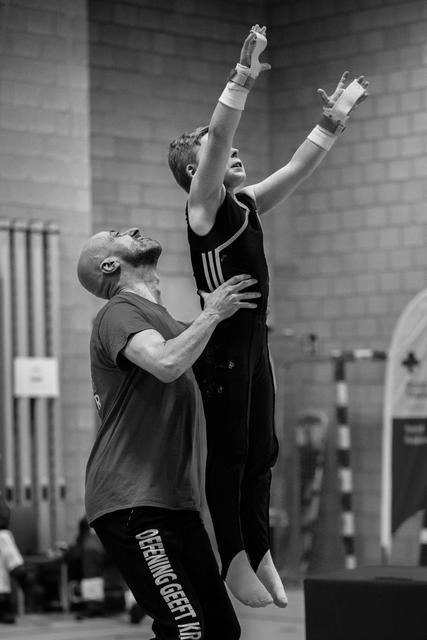 Coach supports gymnast from below as she raises her arms triumphantly, celebrating a successful routine in the gym