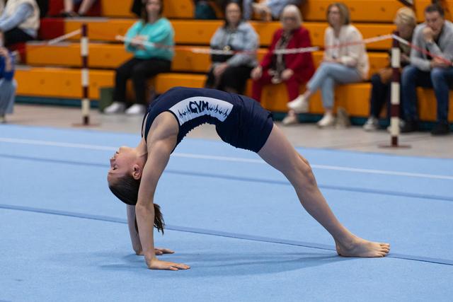 Gymnast performs a dramatic backbend on blue mat while spectators watch from bleachers in gymnasium
