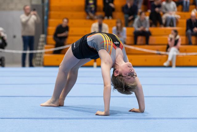 Young gymnast performs a dramatic backbend during floor routine, arching backward with hands planted on blue mat