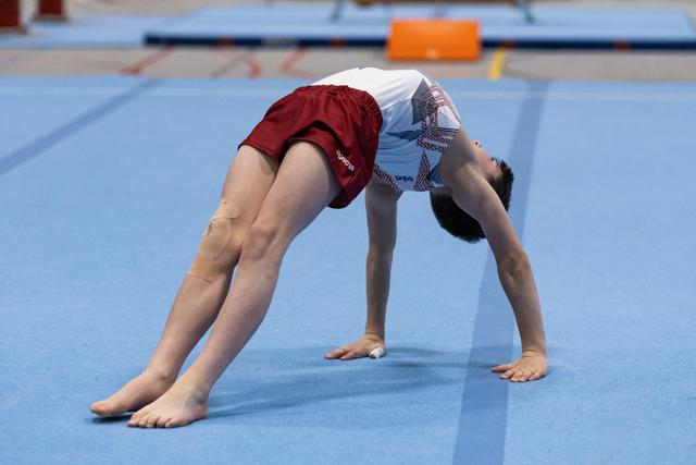 Gymnast in burgundy shorts and light blue leotard performs backbend stretch on blue training mat during warmup