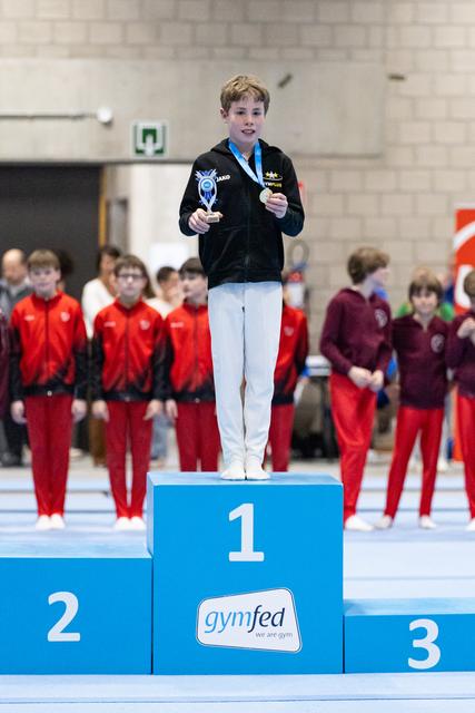 Young gymnast stands proudly on first place podium holding trophy and medal, with competitors in red tracksuits behind