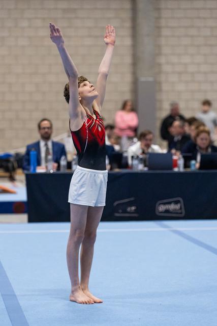 Young gymnast completes floor routine with arms raised upward, displaying focused expression in competition hall