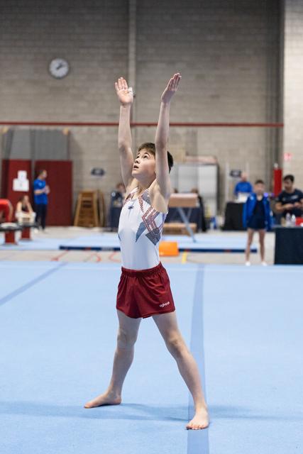 Young gymnast completes floor routine with arms raised overhead, looking up with focused expression in training facility