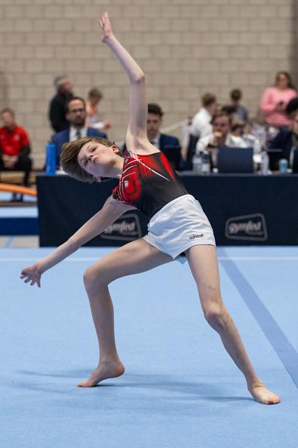 Young gymnast performs an expressive backbend during floor routine, one arm extended upward, at indoor meet with spectators