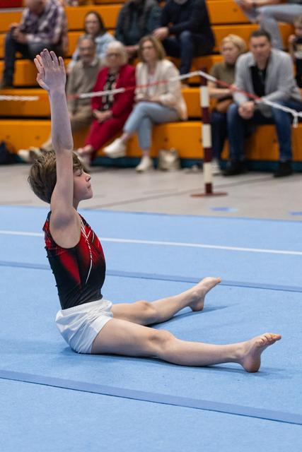 Young gymnast performs a seated floor routine with arms raised, demonstrating flexibility before an audience in the bleachers