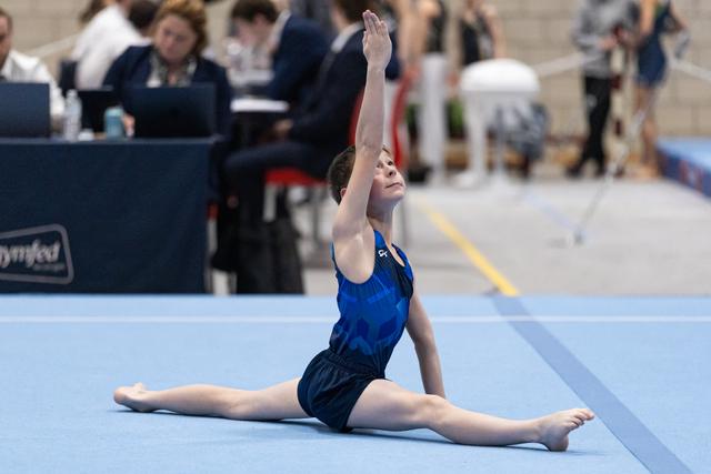 Young gymnast performs a seated split with raised arm during floor routine at indoor gymnastics event
