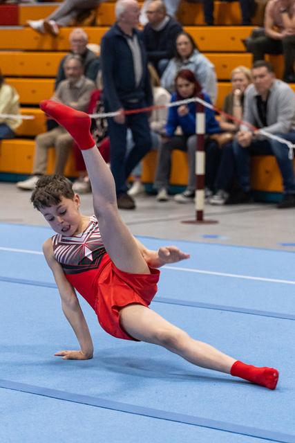 Young gymnast performs a dramatic floor routine in red and white striped leotard, executing a split while spectators watch