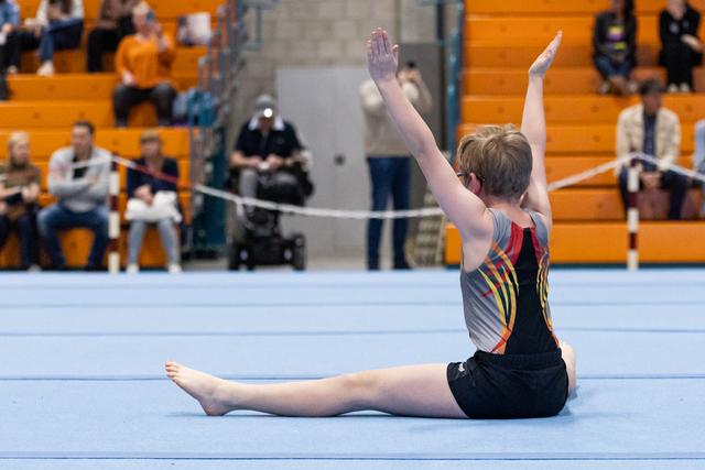 Young gymnast performs a straddle split with raised arms during floor routine at indoor gymnasium meet