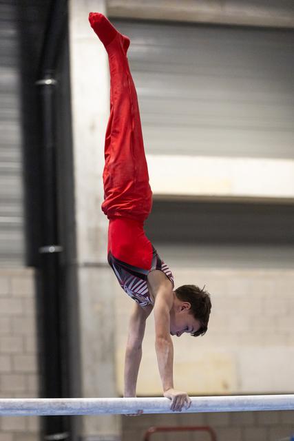 Male gymnast performing a handstand on the balance beam, legs extended vertically in red pants during training