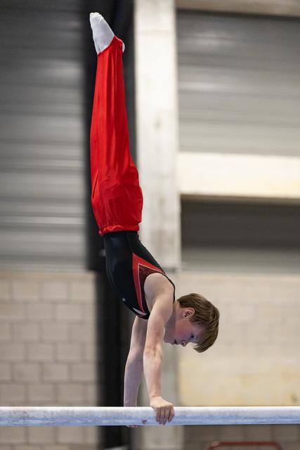 Young male gymnast performs inverted handstand on parallel bars, displaying strength and balance in red and black uniform
