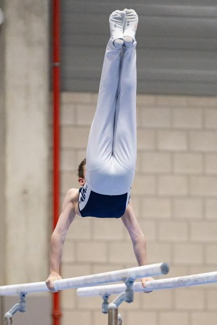 Male gymnast performs inverted handstand on parallel bars, body fully extended upward in white and blue uniform