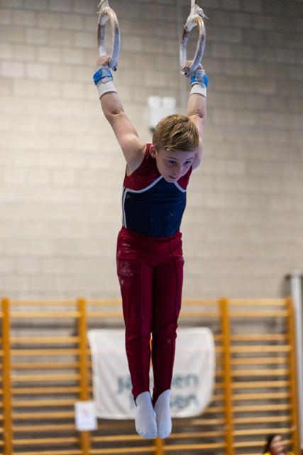 Young gymnast hangs from rings with arms extended overhead, focused expression, wearing burgundy and navy uniform in gymnasium