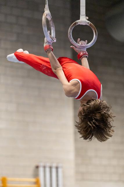 Gymnast performing an inverted hold on still rings, body horizontal in bright red uniform with white trim