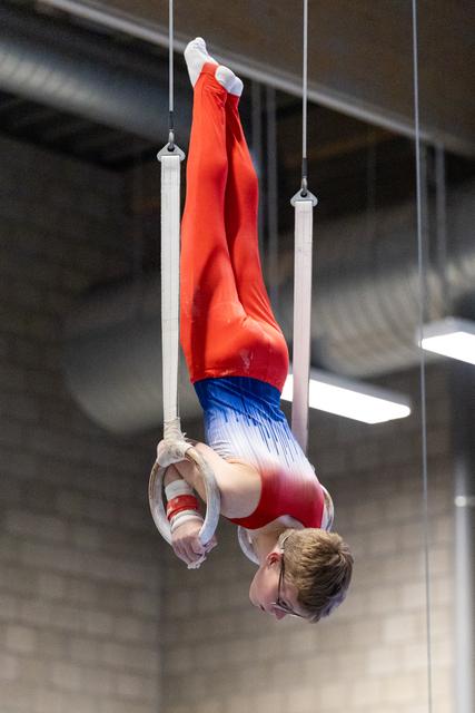 Gymnast performs inverted hang position on still rings, demonstrating strength and control in red and blue uniform