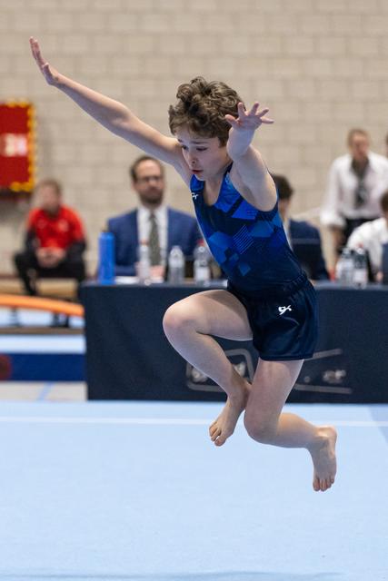 Young gymnast performs an expressive floor routine, suspended mid-air with arms raised and knees bent during her tumbling pass.