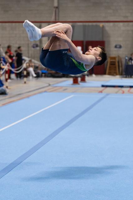 Gymnast performs a tucked backflip during floor exercise routine on blue mat in indoor training facility