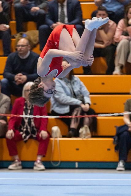 Gymnast performs inverted backflip above floor mat with spectators seated on yellow bleachers in background
