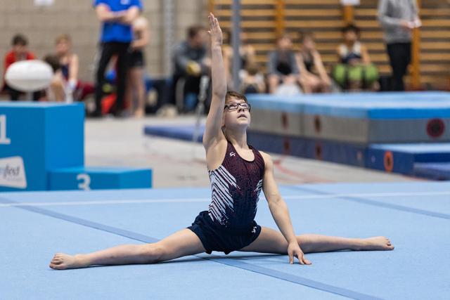 Young gymnast in sequined leotard performs graceful split pose on floor mat, one arm extended upward during routine