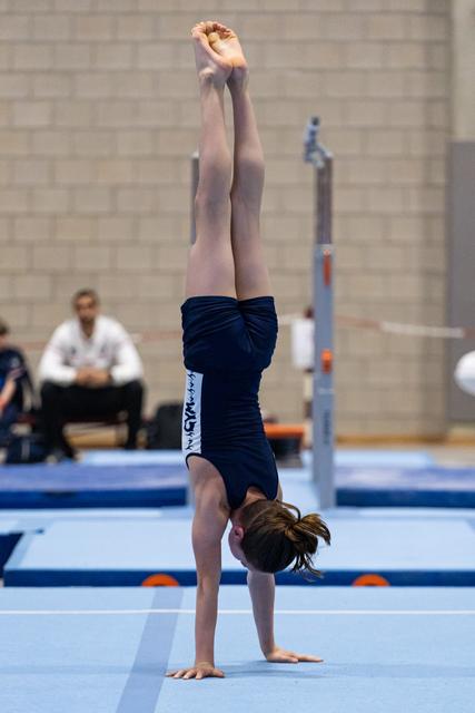 Young gymnast executes a controlled handstand on blue floor mat during routine, coaches observing in background