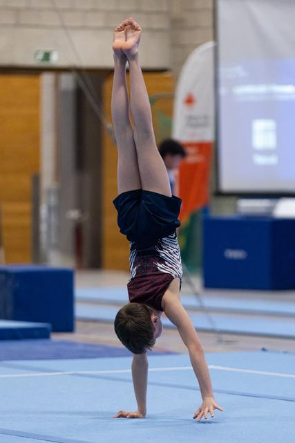 Young gymnast executes a vertical handstand on blue floor mat, legs extended upward with pointed toes in training facility