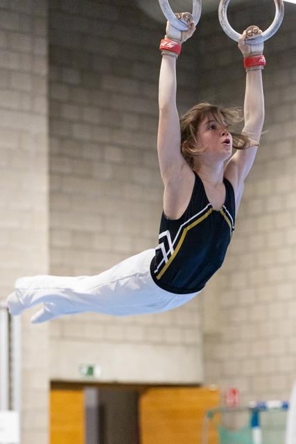 Young gymnast performs a horizontal hold on still rings with focused expression, wearing navy leotard in training facility