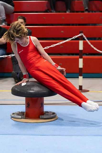 Gymnast in red uniform performing pommel horse routine with focused expression, body extended horizontally in gym