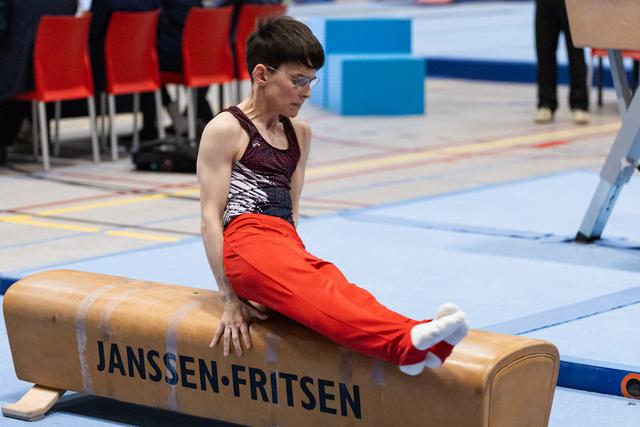 Male gymnast in sequined leotard and red pants sits on pommel horse, focused and preparing for his routine at indoor meet