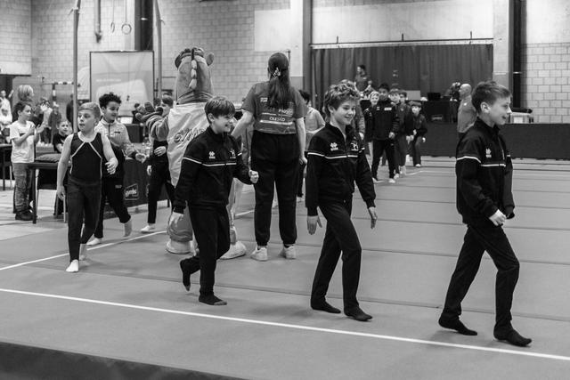 Young gymnasts in black tracksuits march in formation across a floor exercise mat during an opening ceremony at an indoor venue