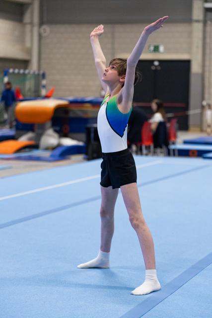 Male gymnast performs floor routine with arms gracefully extended upward, wearing blue and white leotard in training facility