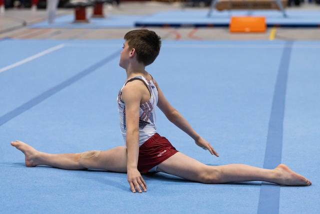 Male gymnast performs a floor split stretch on the mat, demonstrating flexibility during warm-up at a gymnastics venue.