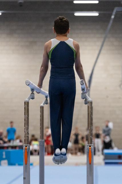 Male gymnast in navy and green leotard executes support hold on parallel bars during routine at indoor gymnastics meet