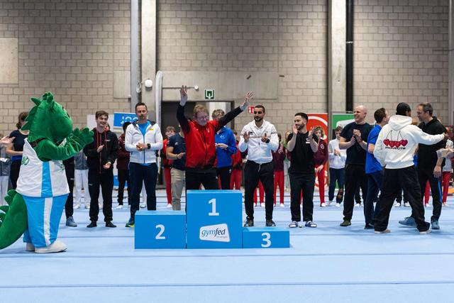 Gymnast in red celebrates triumphantly on first place podium while crowd applauds, with green mascot visible at left