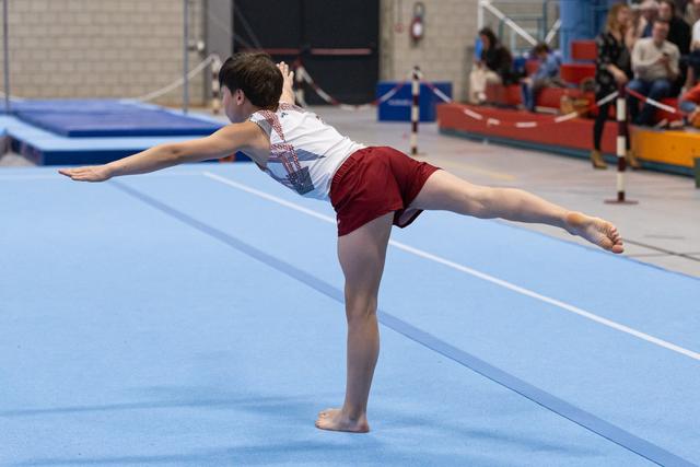Young gymnast holds an arabesque balance on blue floor mat, arms extended gracefully during floor exercise routine